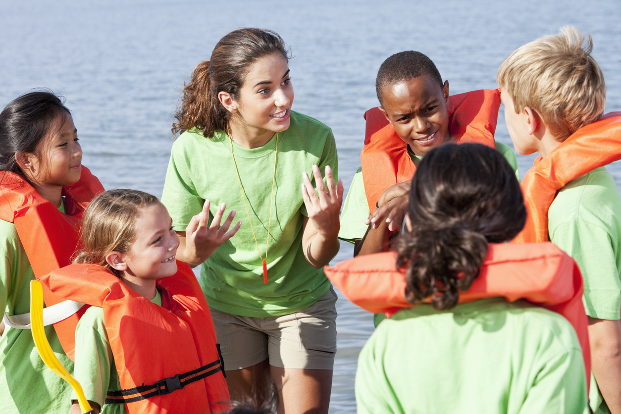 Teenage girl talking with group of children wearing life vest by lake.