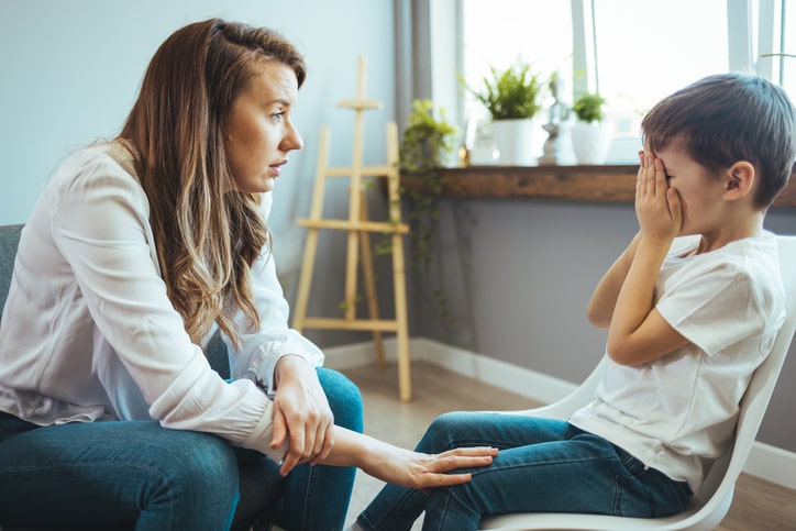 Upset little boy crying in psychologist's office unable to control emotions, sharing problems and traumas, professional psychotherapist comforting kid, side view