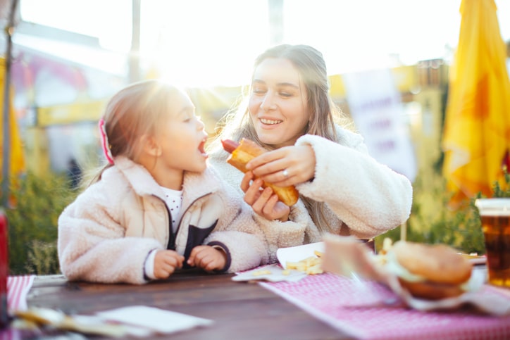 Mother and daughter eat hotdog lunch outside on picnic tables in the sun