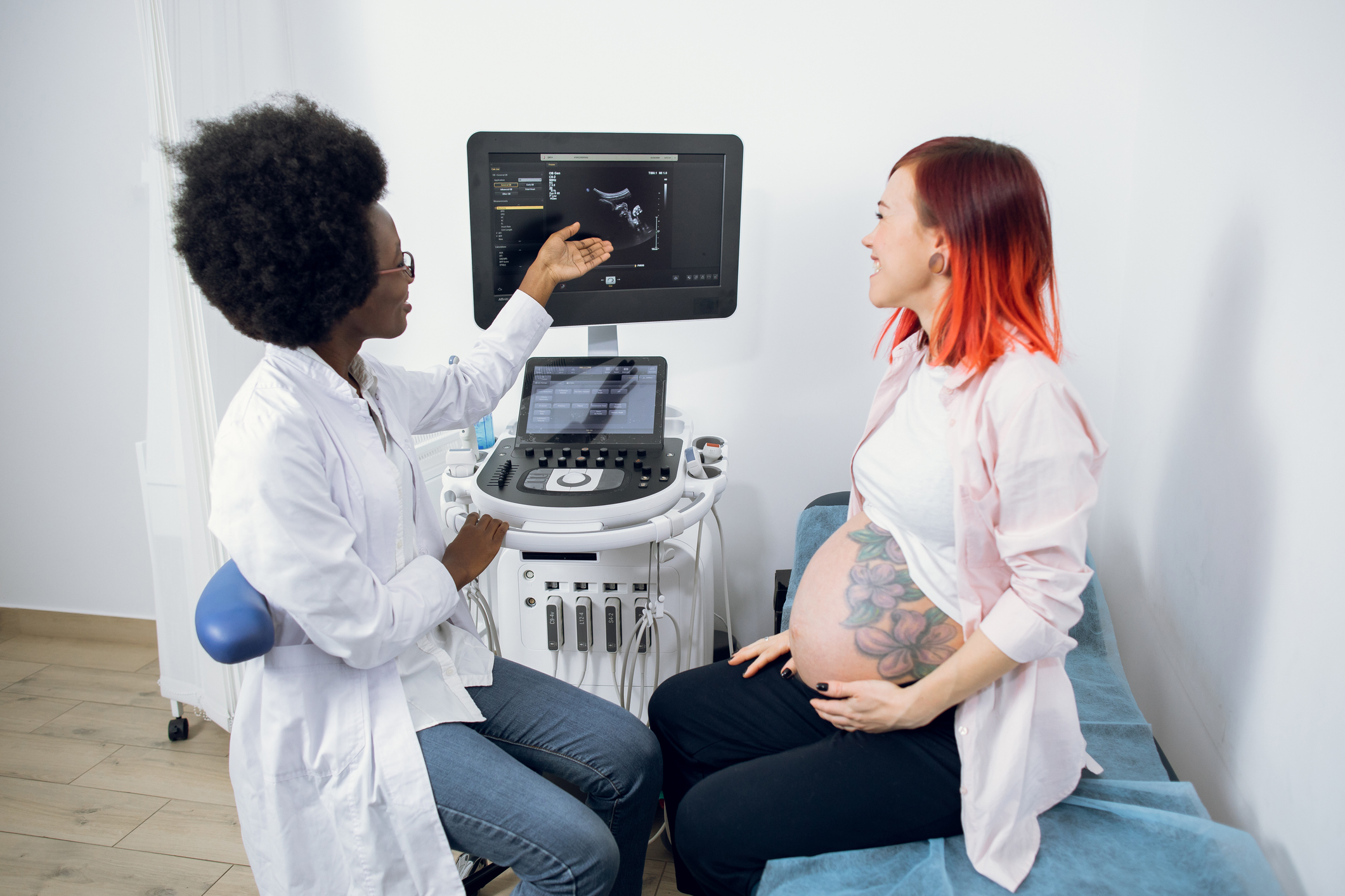 Beautiful young red haired pregnant woman in modern clinic, looking at the screen of ultrasound machine and asking her female African doctor obstetrician about the results of ultrasound test