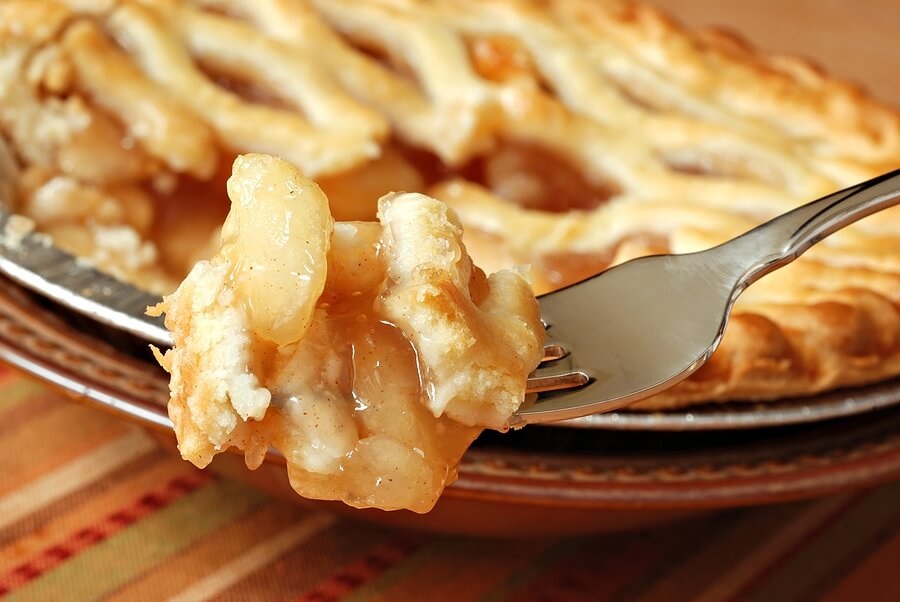 Mouthful of apple pie on fork, with pie in background.