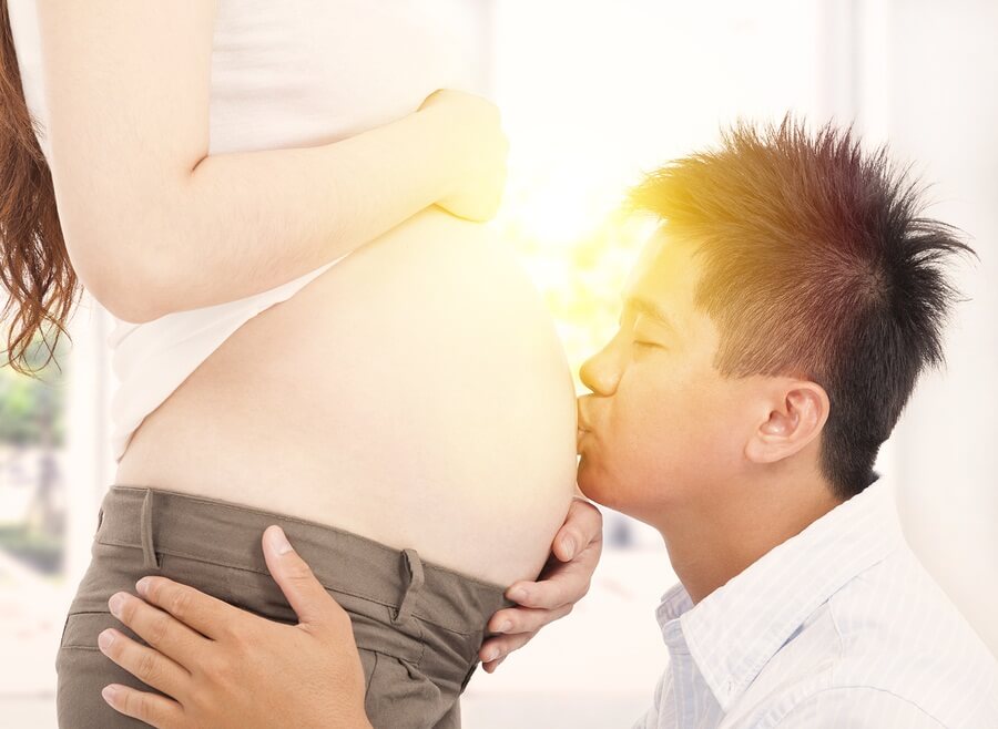 Asian man kissing pregnant belly with sun in background