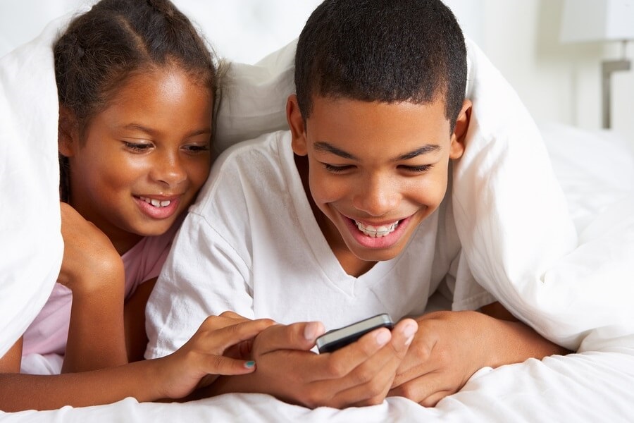 African American brother and sister using smartphone under duvet