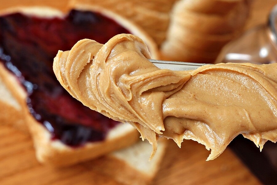 Close up of peanut butter on knife with bread and jam in background