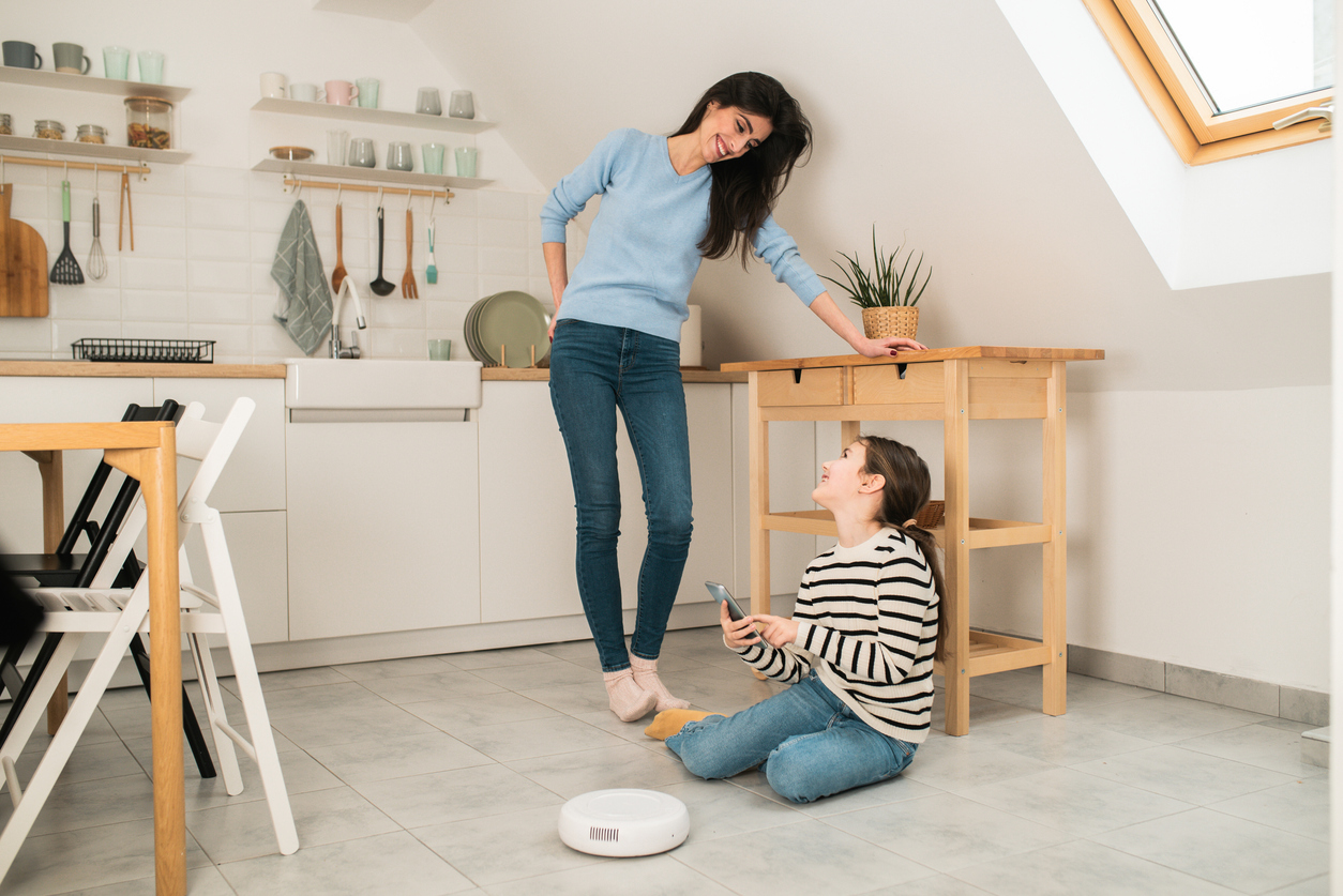 Young woman and her little daughter turning on a robot vacuum cleaner using a smart app on the smartphone