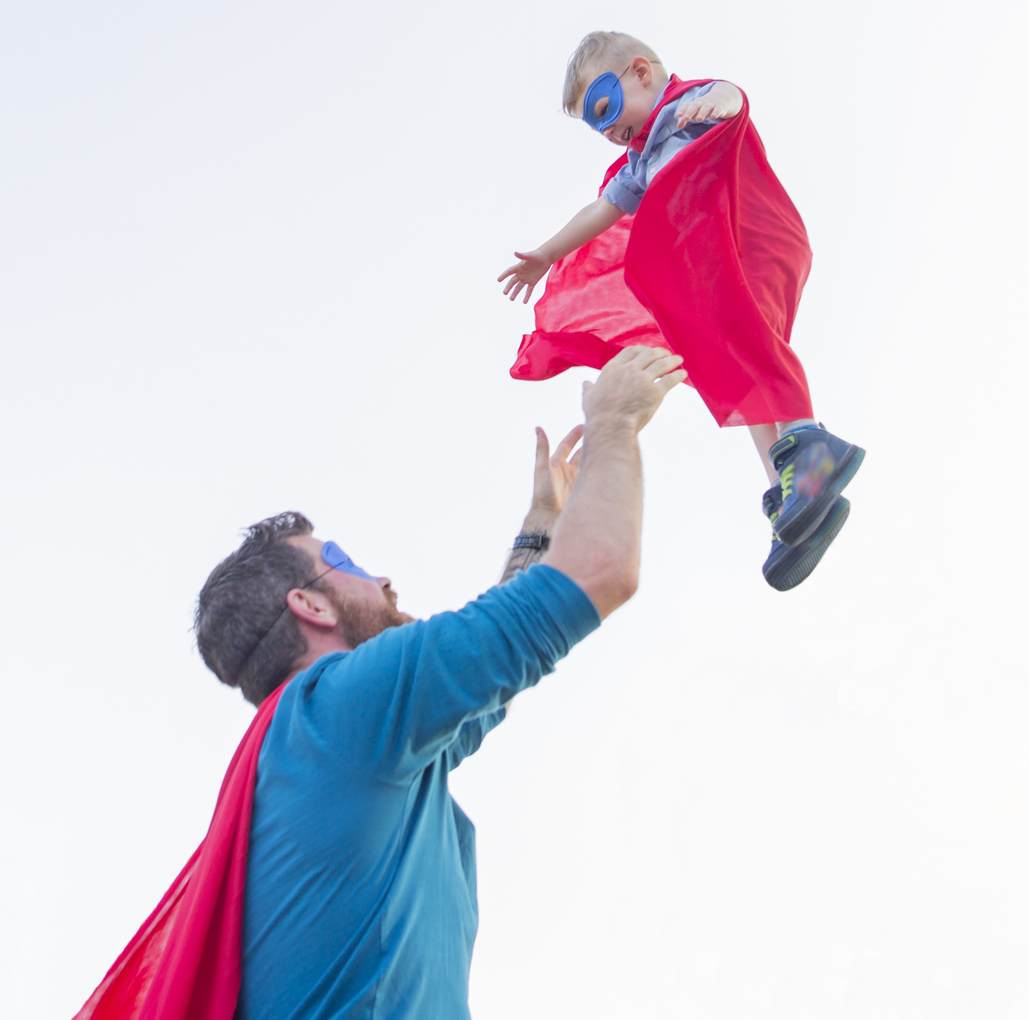 Cute little three year old boy wearing a superhero outfit laughing as he is being thrown in the air playfully by father, who is also wearing a red cape and blue mask.