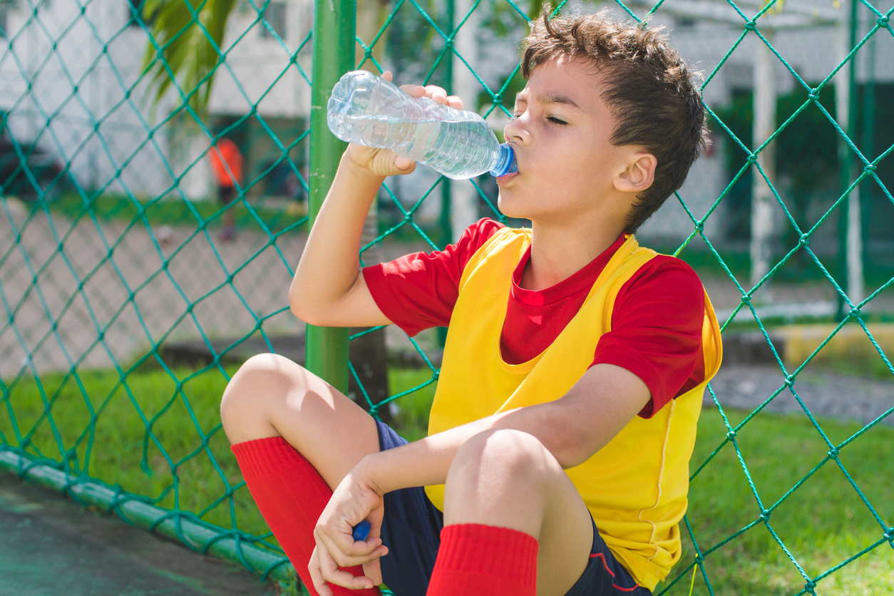 Exhausted young boy drinks water out of plastic bottle after playing soccer