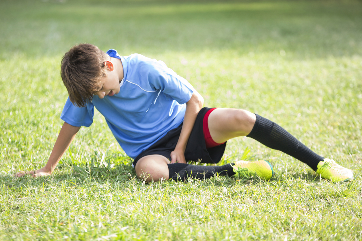  teenage boy holds his leg while grimmacing in pain after sustaining injury during soccer game.