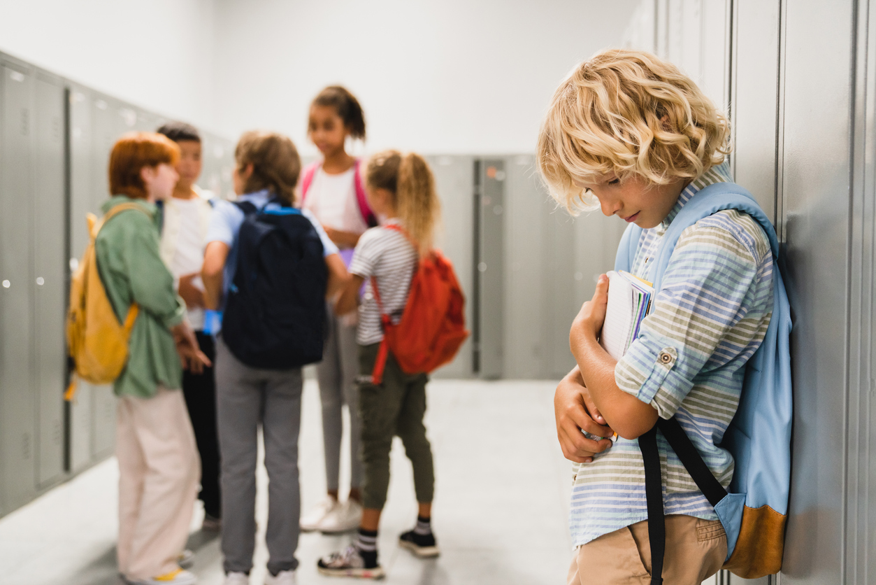 elementary school child stands alone at his locker, excluded from group of friends in background