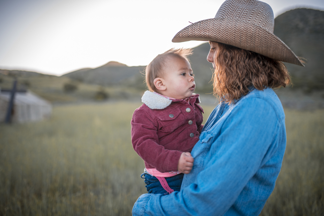 Mother holds baby in field. Mother wears cowboy hat.