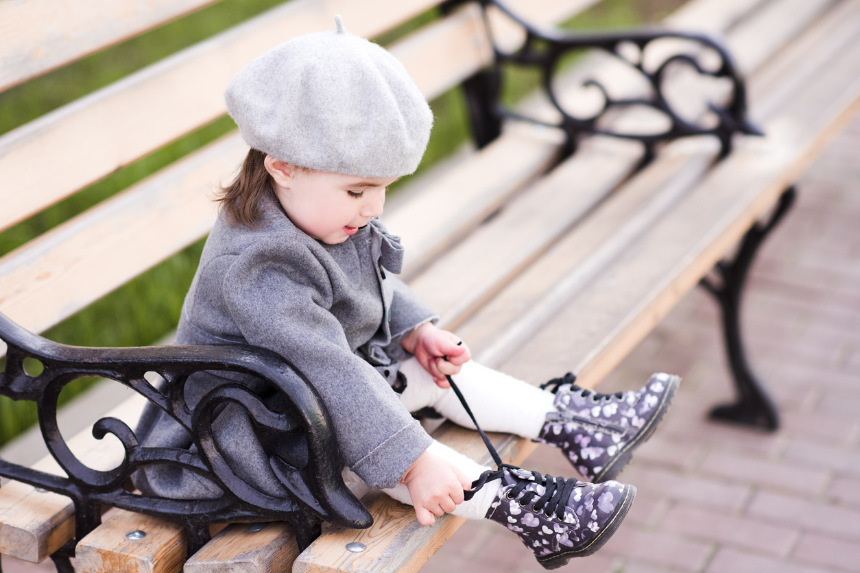 Baby girl lace shoes sitting on bench in park. Wearing french style beret and coat. Childhood.