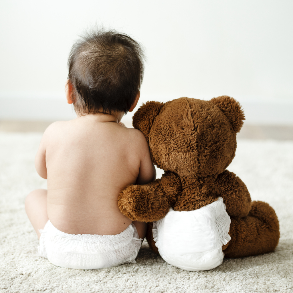 Back of a baby with a teddy bear. Both the baby and bear wear diapers.