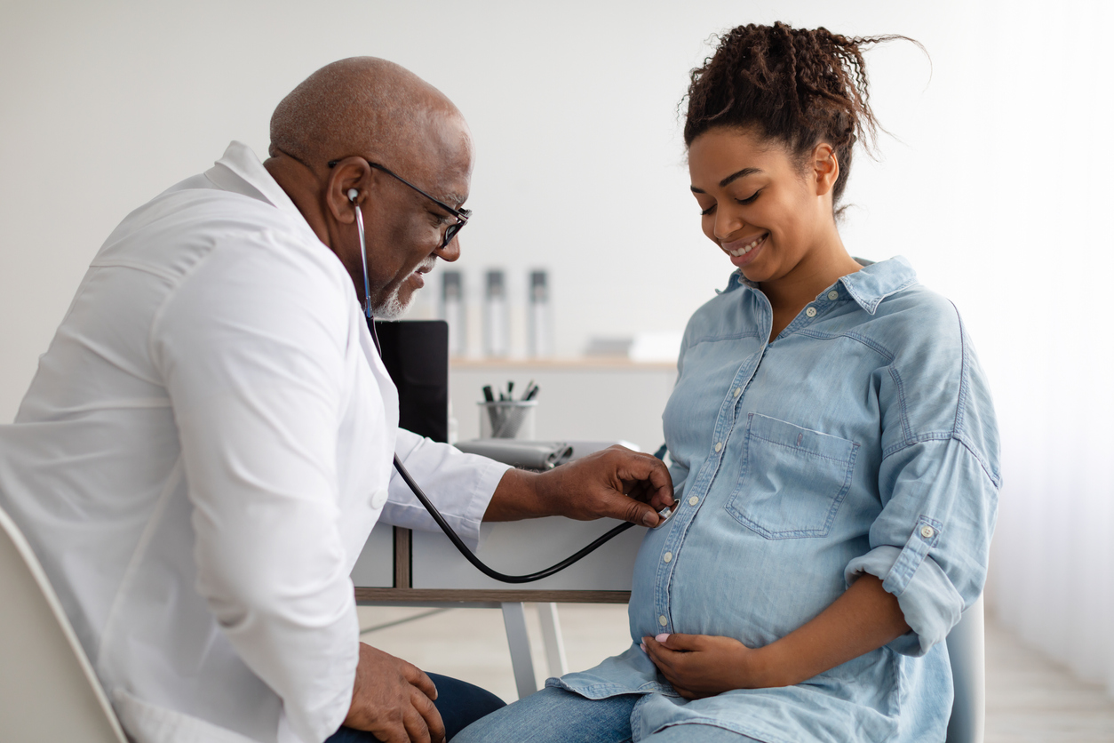 Gynecology Consultation. Smiling black pregnant woman visiting her obstetrician doctor in maternity clinic, mature male gynaecologist examining her belly with stethoscope, doing medical check-up