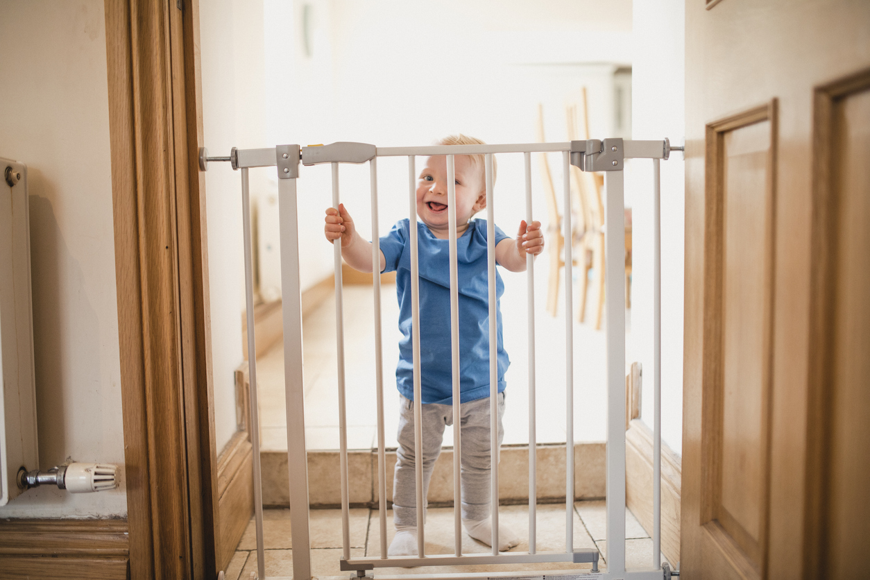 A joyful child smiles whilst holding on to a locked safety gate at home, he is wearing casual clothing and has a big grin on his face.