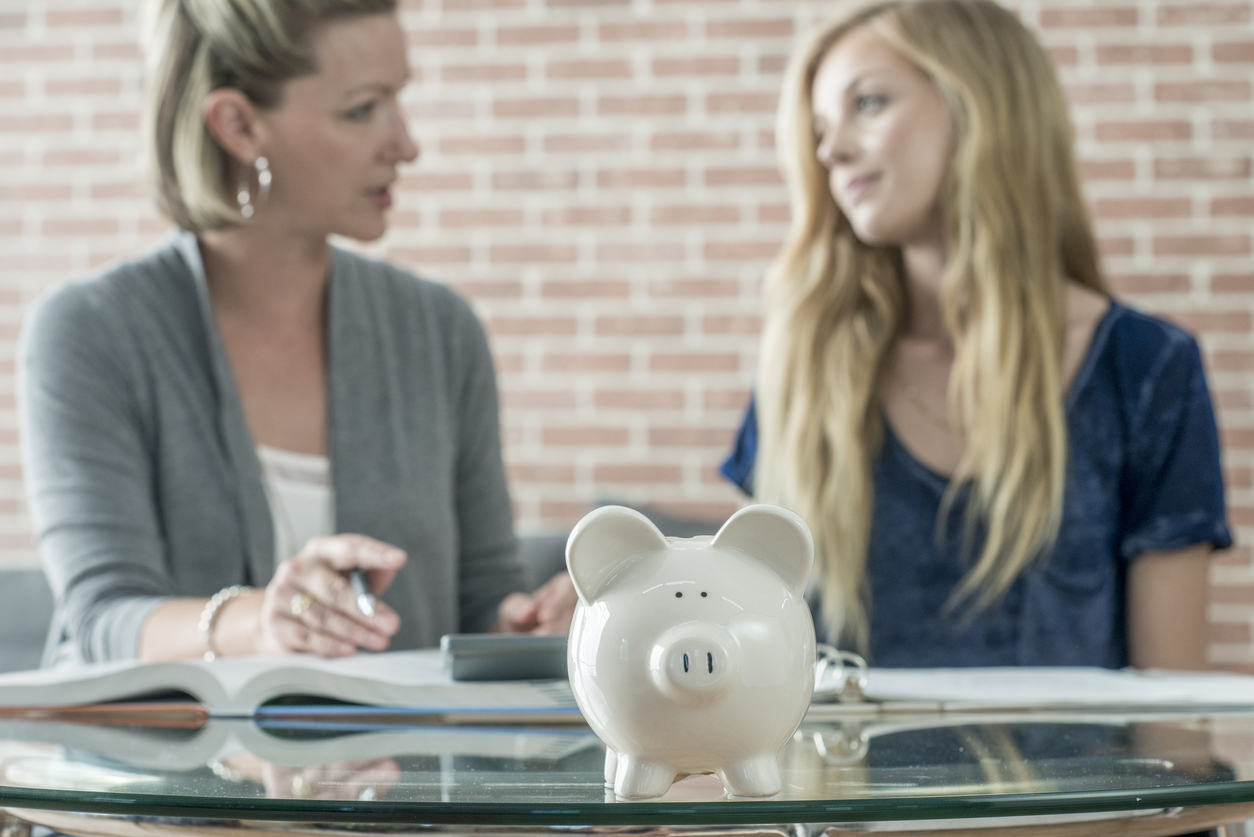A businesswoman and tutor is teaching a high school student the basics of finances, savings, bank accounts, credit cards, loans, and debt. A piggy bank is sitting on the table.