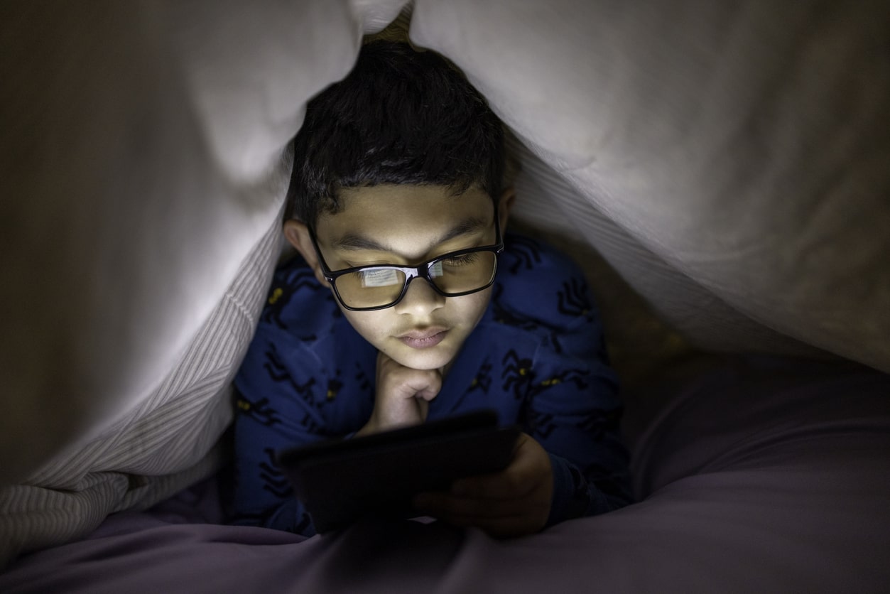 Young boy sits on iPad under the covers of his bed