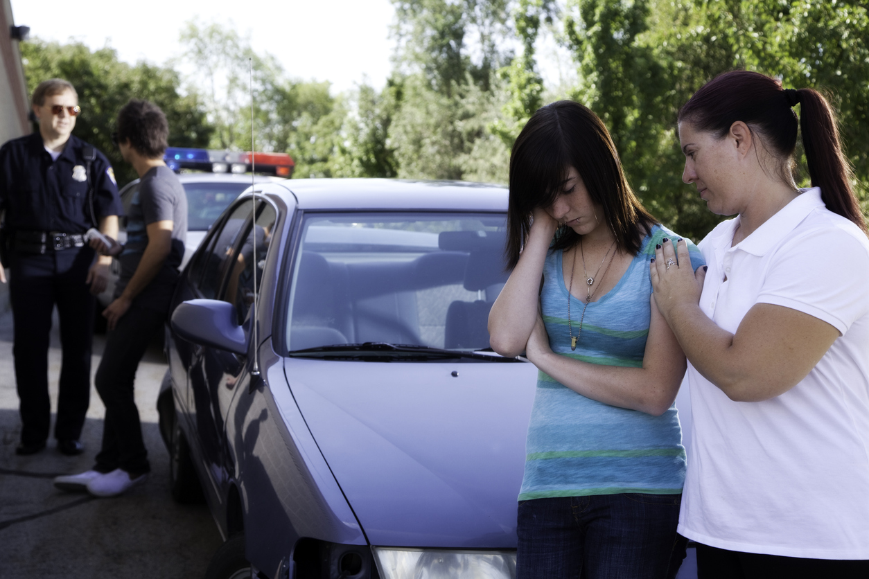 A mother comforts her daughter after a minor accident or traffic stop, which the police officer talks to her boyfriend in the background