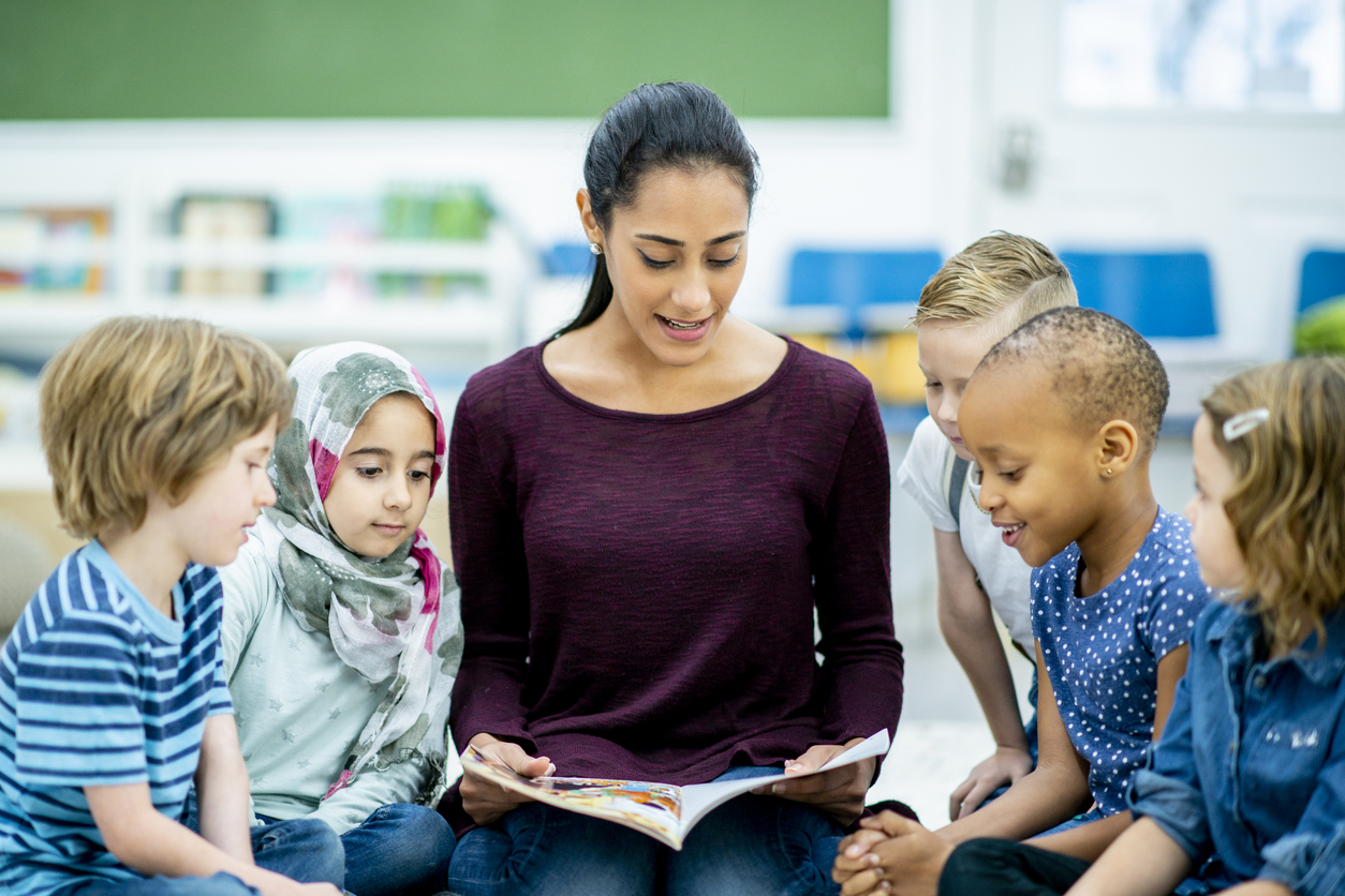 A diverse group of kids gathers around a female teacher of Middle Eastern descent and listens attentively to the story she is reading.