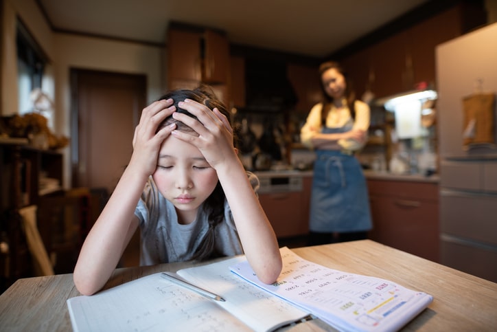Stressed young girl struggles over her homework, while mother watches in the background. Adaptogens and Stress.