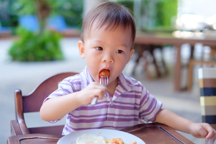 Toddler boy child sitting in high chair using fork eating whole cherry tomatoes