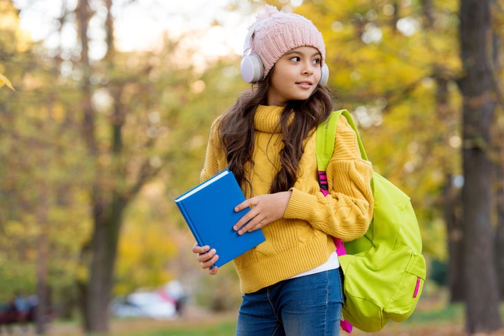 kid with book and backpack listens music in earphones, school.