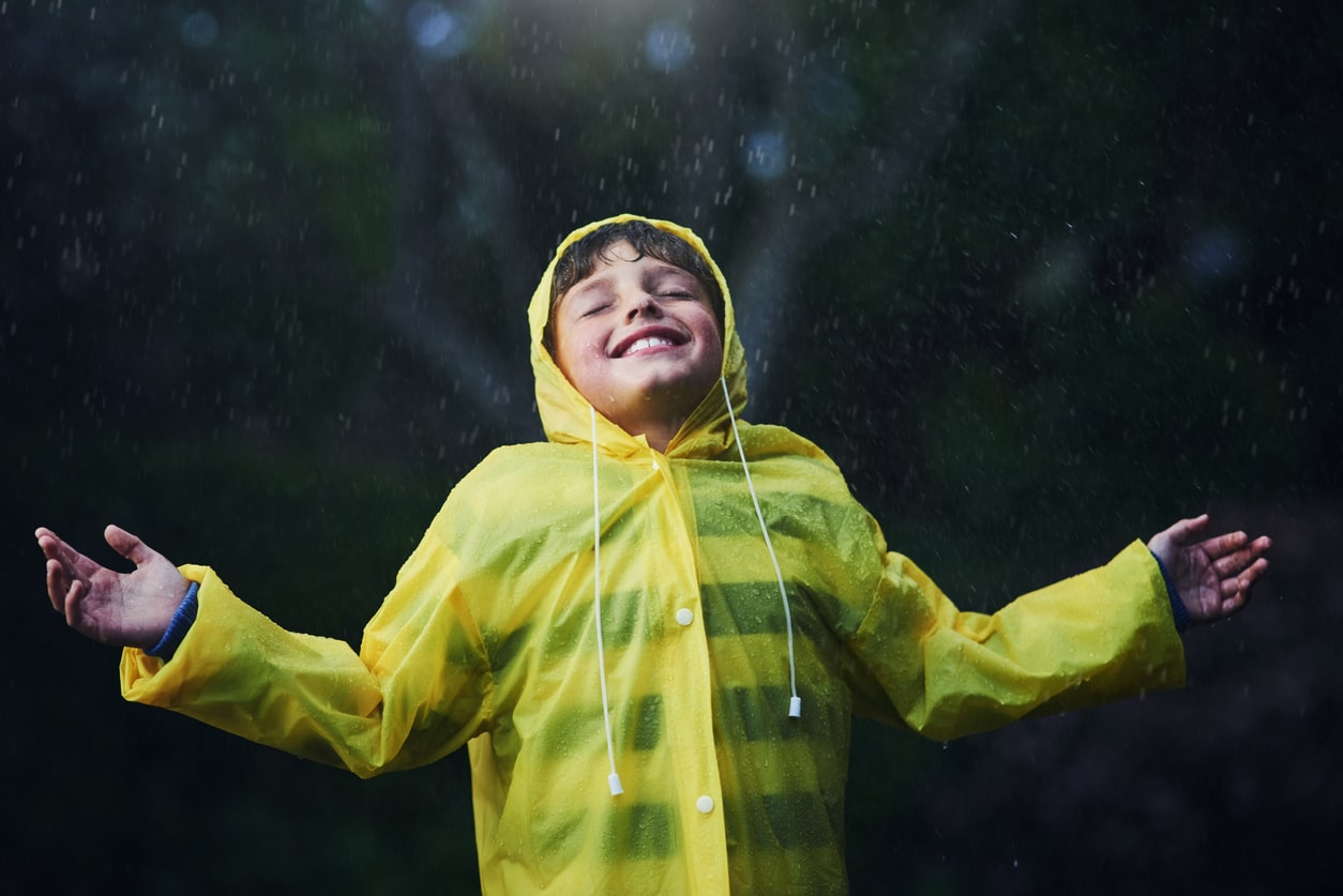 Young boy stands outside in the rain with a big smile. He wears a yellow rain jacket.