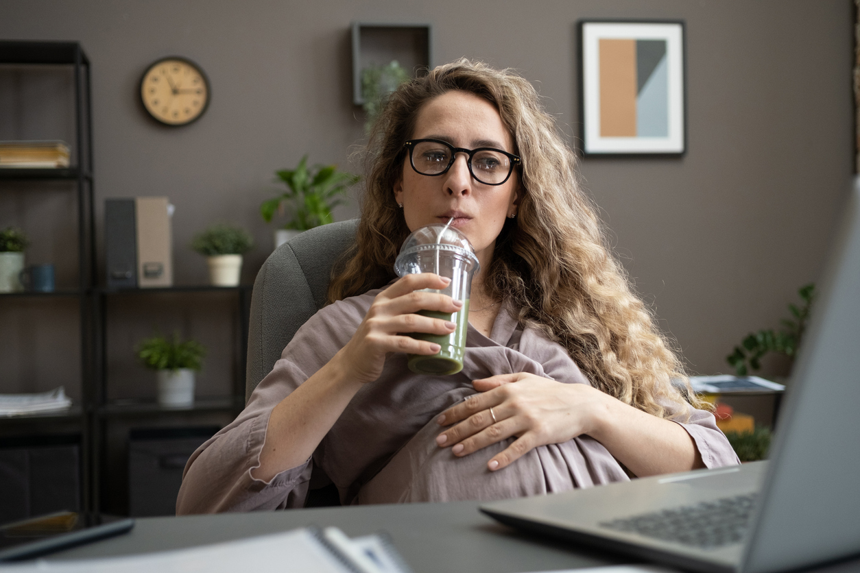 Pregnant businesswoman drinking smoothie while sitting by desk in front of laptop
