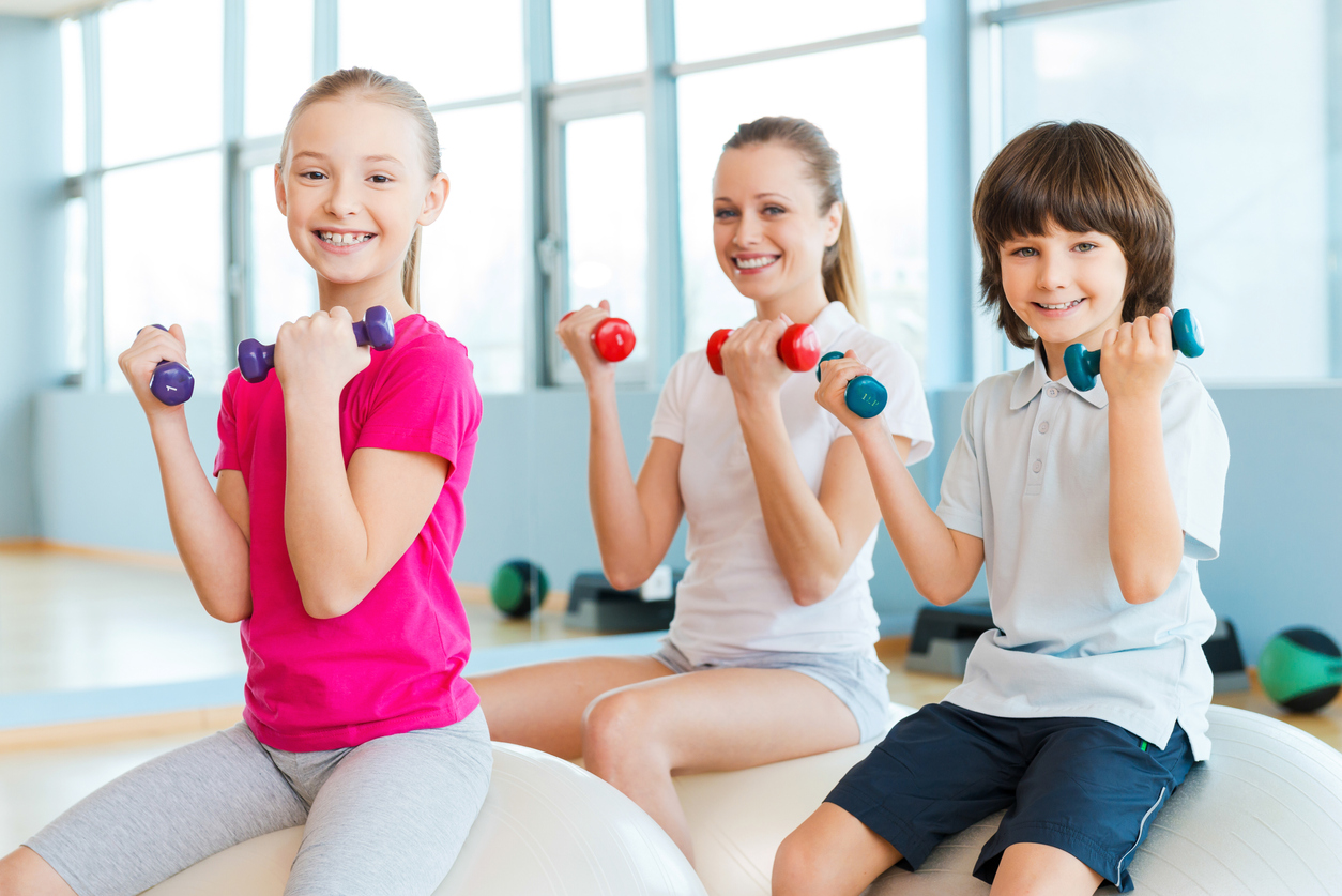 Cheerful mother and two children exercising with dumbbells in health club while sitting on the fitness balls together