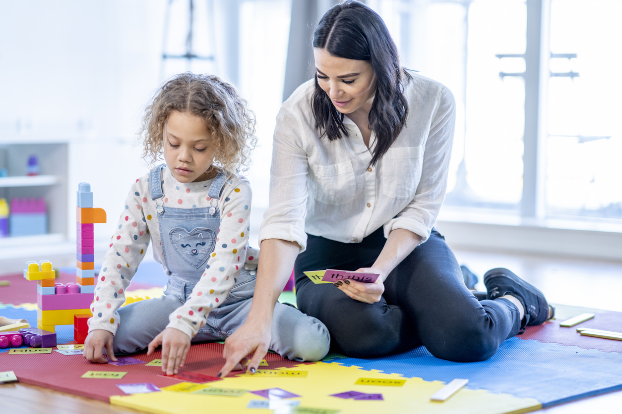 A kindergarten teacher sits on the floor with her young student, as they practice spelling words.