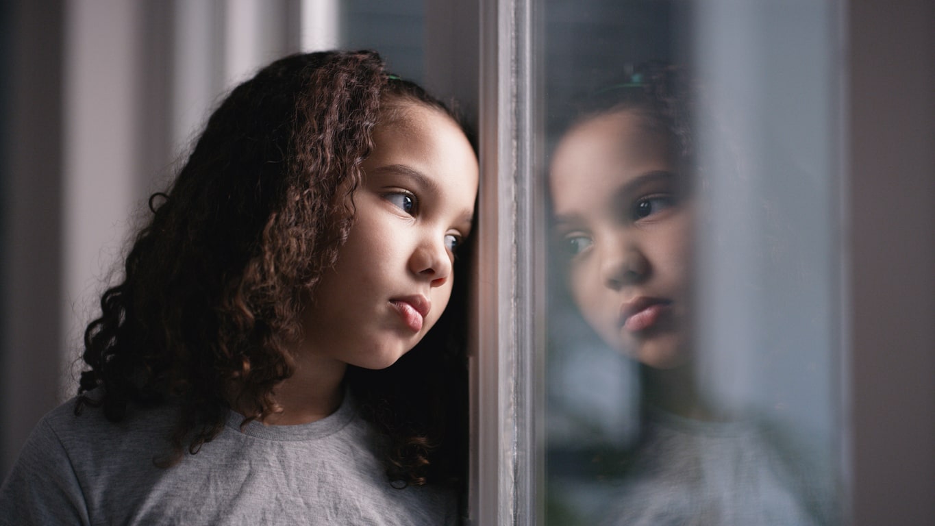 Young girl with curly hair looks sadly out her bedroom window. Concept of generational trauma.