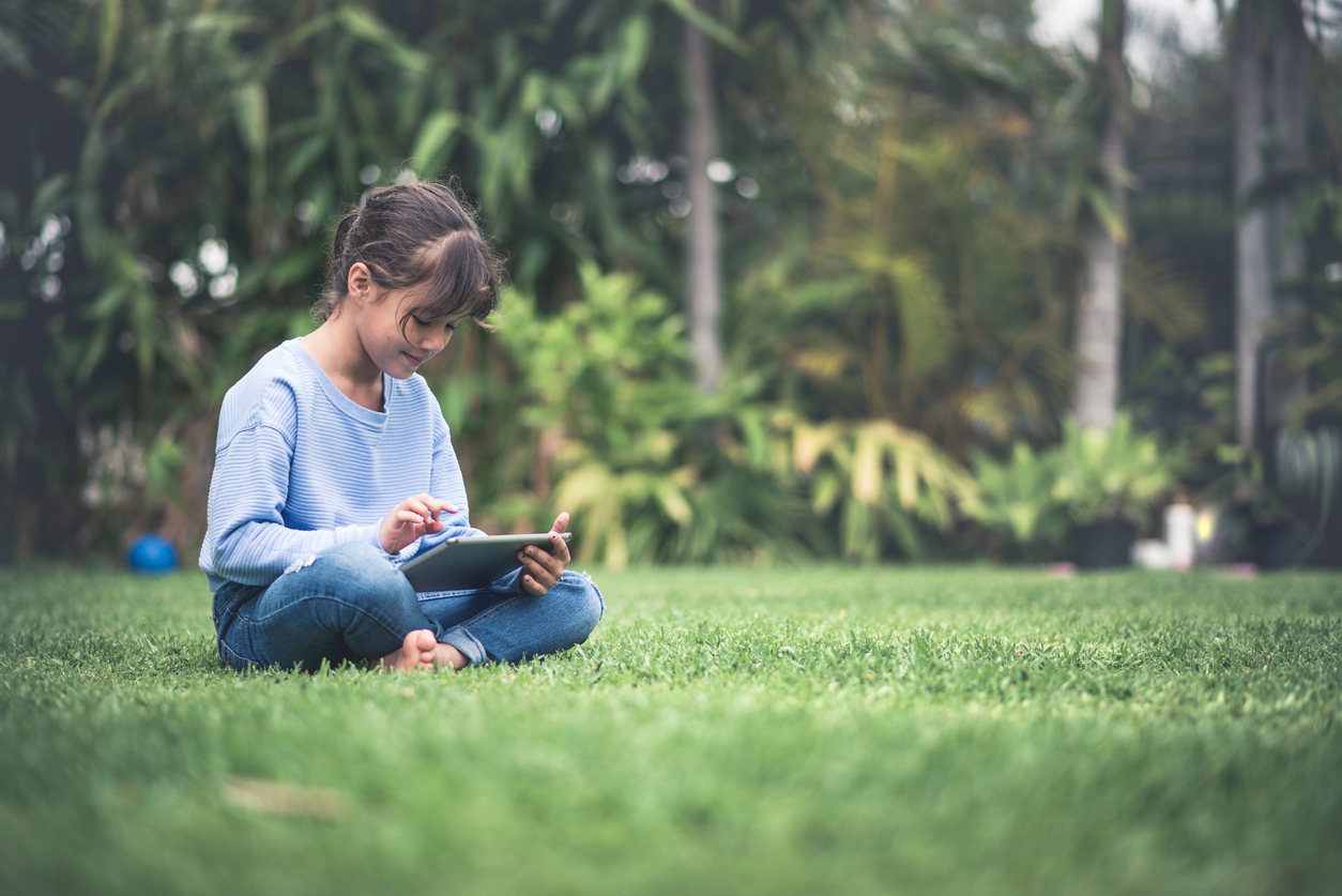Young girl reading on digital tablet. She is sitting on the grass on the backyard, looking at tablet and smiling.