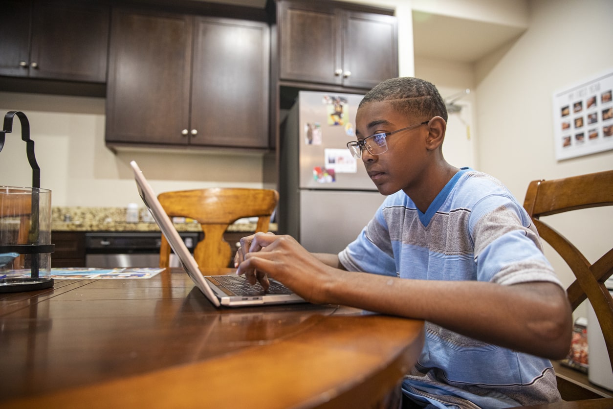 Young boy sits on his laptop in the kitchen. Gen Z falling victim to online holiday scam.