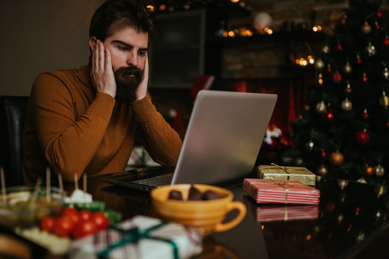 Sad father sits in front at his laptop in front of the Christmas tree. Sad image of holiday scam victim.