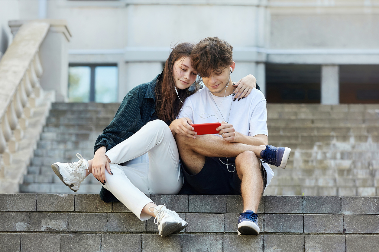 young guy and a girl sitting on the street, who have one pair of headphones for two, look into a red smartphone with a smile.