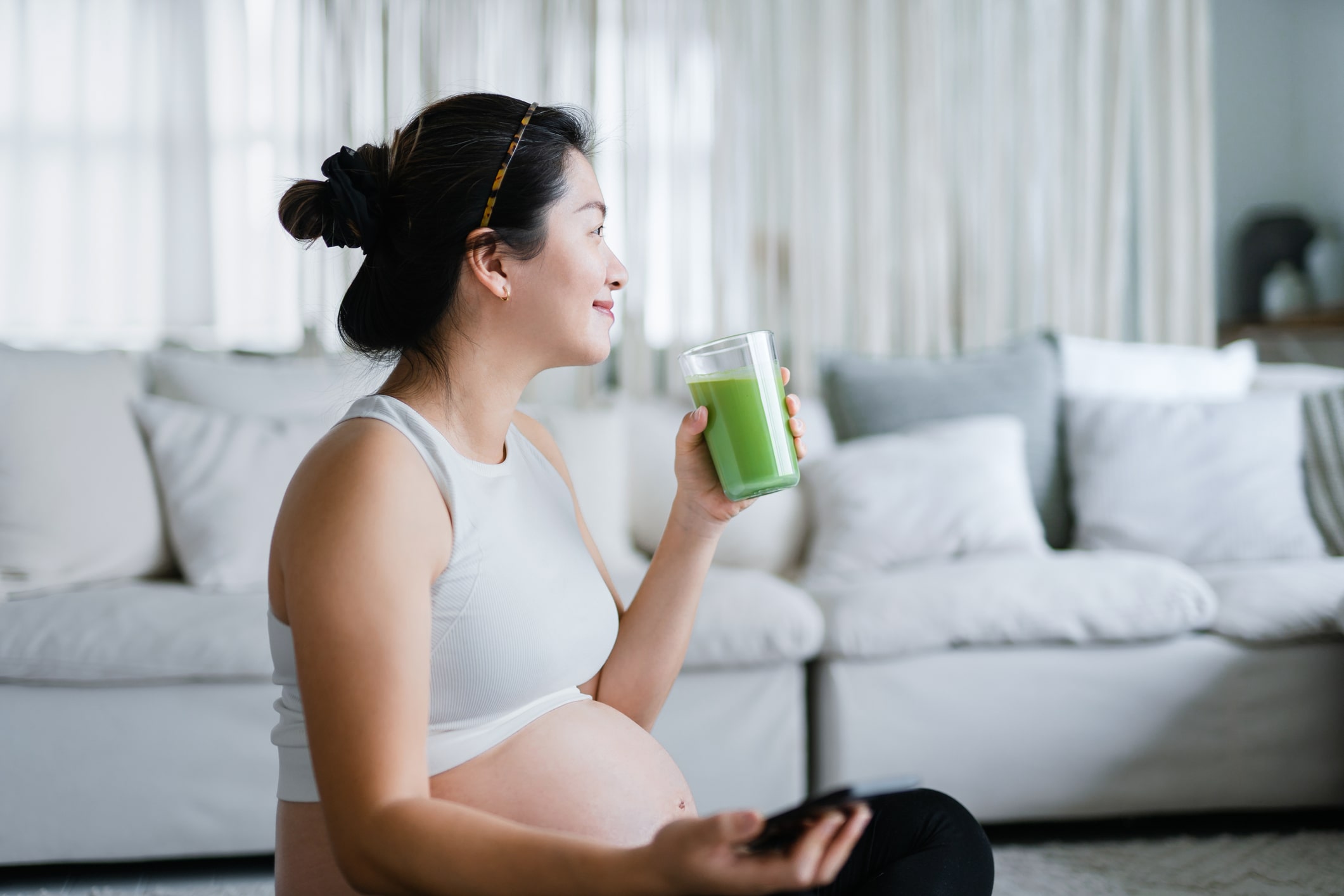 Cheerful young Asian pregnant woman taking a rest after exercising at home, having a glass of fresh home-made healthy green juice. Wellbeing, staying fit and healthy during pregnancy