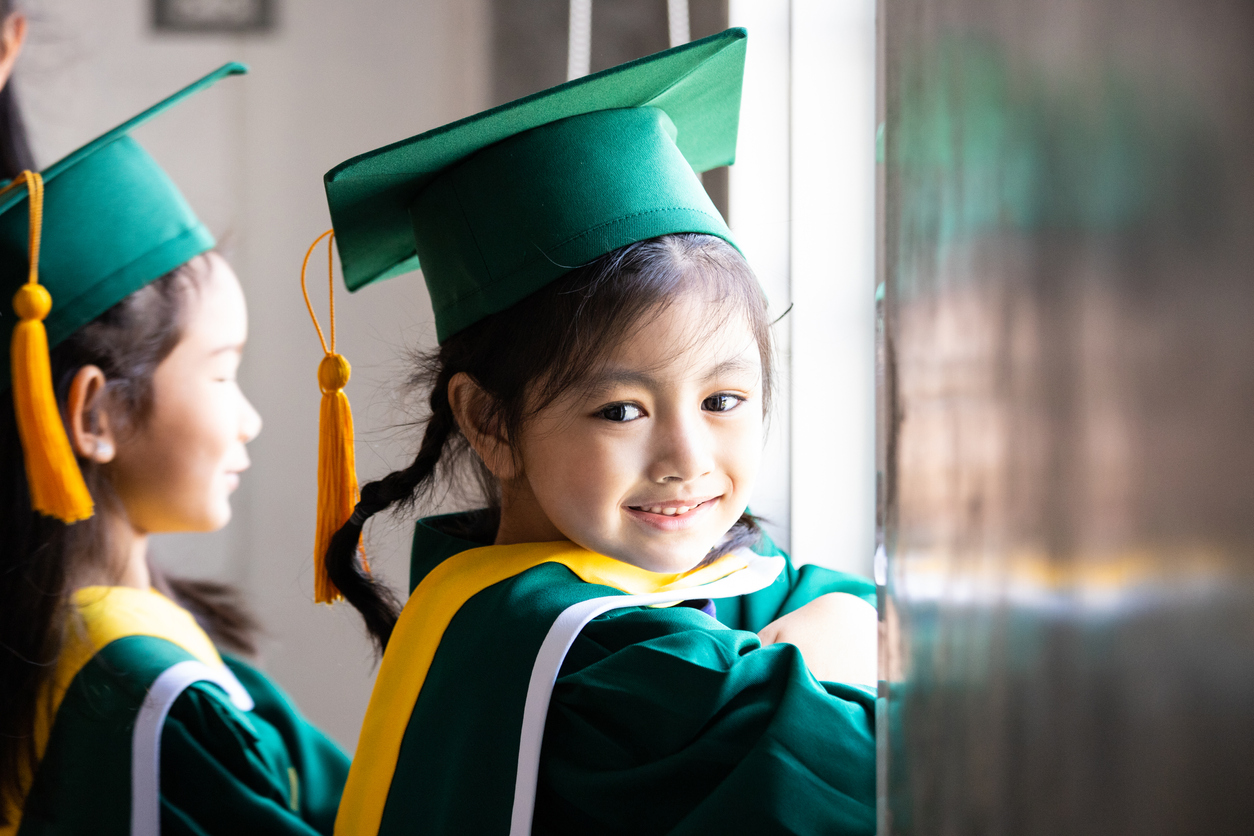 elementary age girls wearing graduation caps and gowns smile while standing inside school.