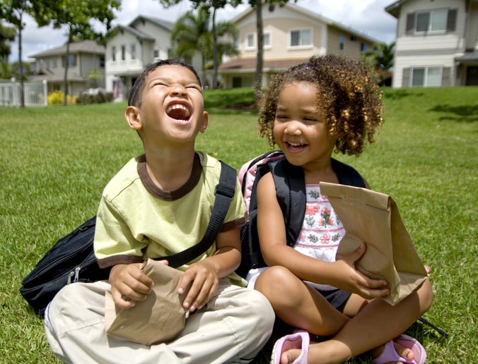 Two young kids sit together outside holding paper lunchboxes