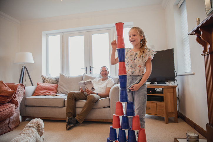 A child stacks cups in the living room whilst her Father reads a book.