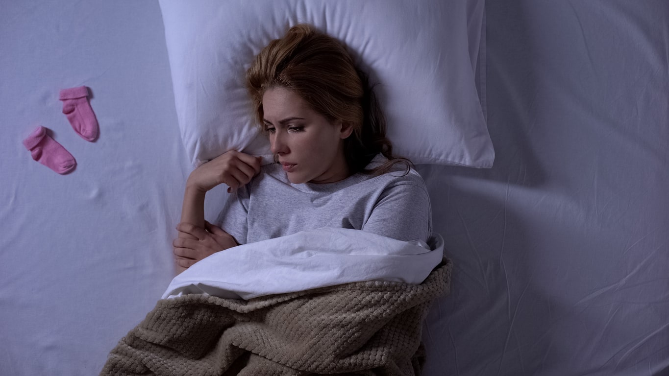 Unhappy woman lying in bed with baby socks beside her