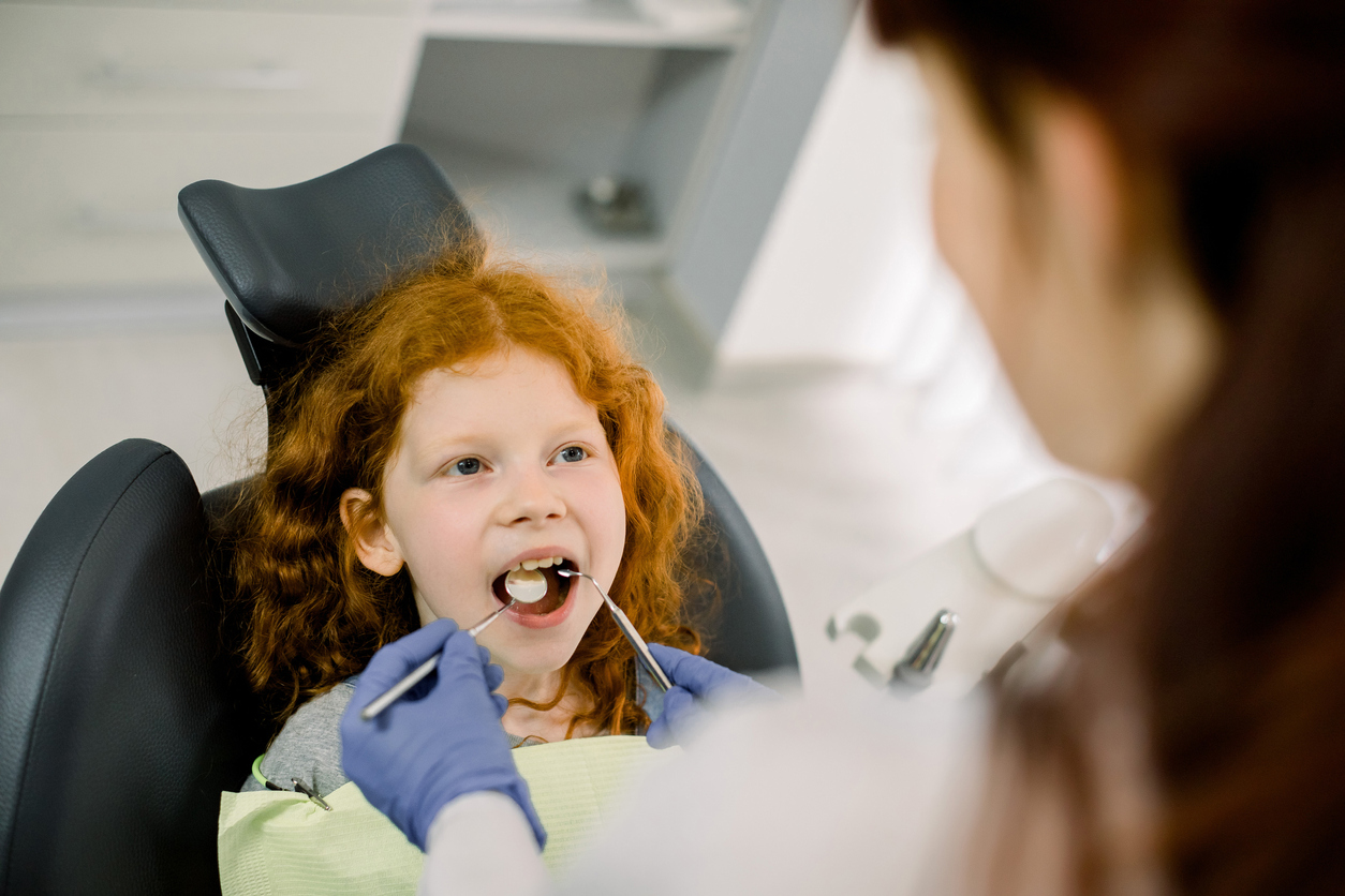 Top back view of female dentist in blue latex gloves checking condition of teeth of little smiling red haired curly girl, sitting in dental chair in modern dentistry clinic. Pediatric dentistry.