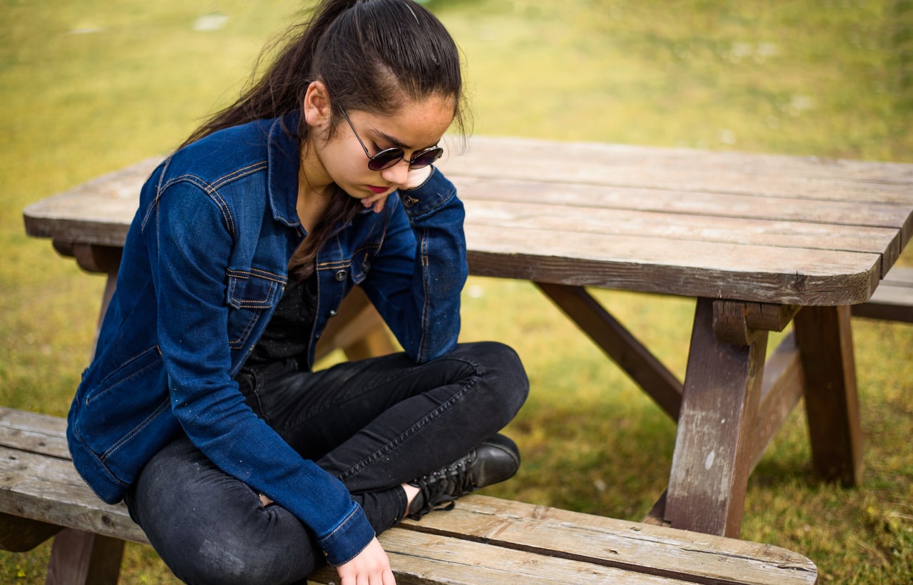 Teenage girl wears sunglasses and sits alone on a park bench, looking sad