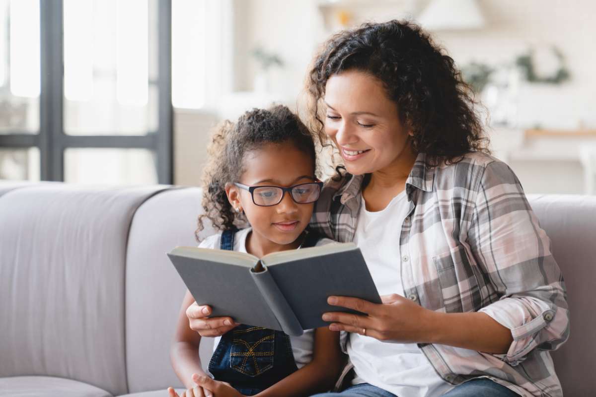 African-american mother mom reading fairy tales book novel with daughter girl, learning together, helping assisting with homework for school. Female tutor teaches schoolgirl at home. Homeschooling